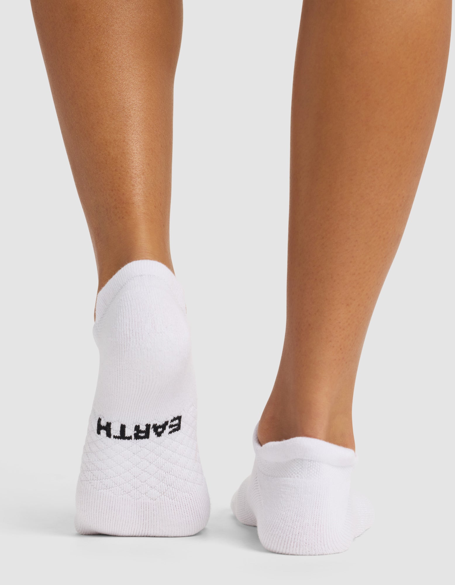 Back view of two feet wearing Cozy Earth Essential Ankle Socks in white. "EARTH" is written in black on the heel of the left sock. The background is plain white.