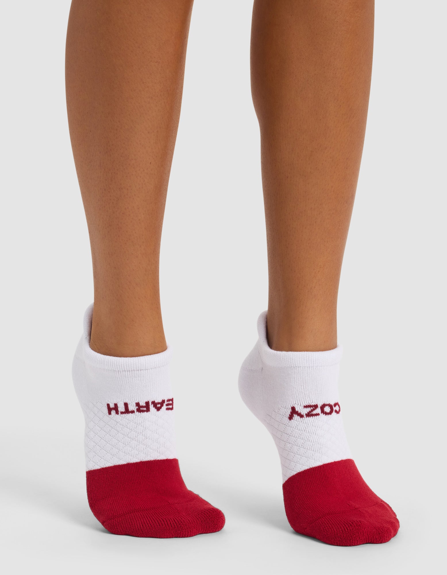 A person wears Cozy Earth Essential Ankle Socks—white with red toes and heels. "EARTH" is printed on the left cuff, while "COZY" appears upside down on the right sock.