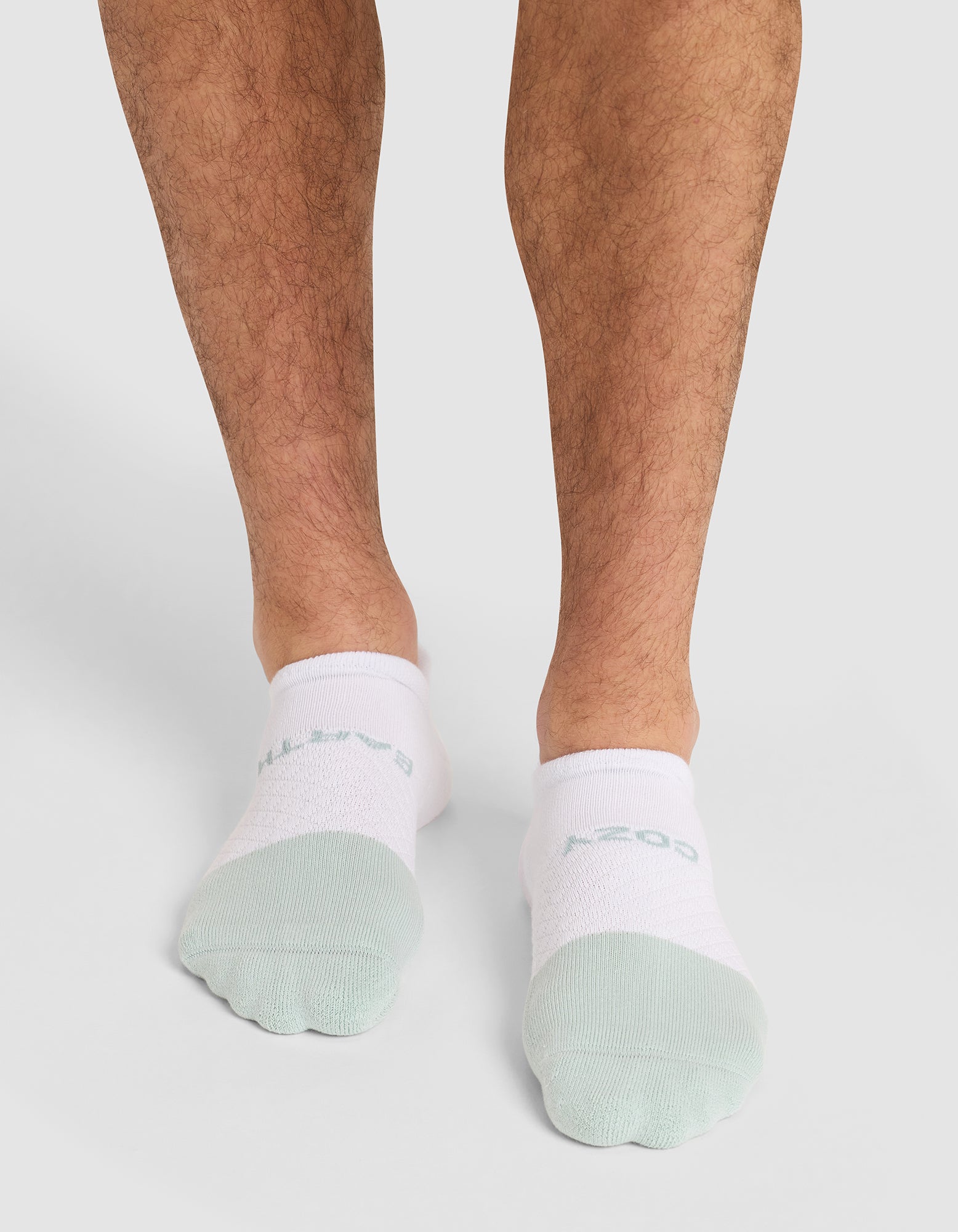 A person wears Cozy Earth Essential Ankle Socks in white and light green, standing on a light background with only their lower legs and feet visible. 