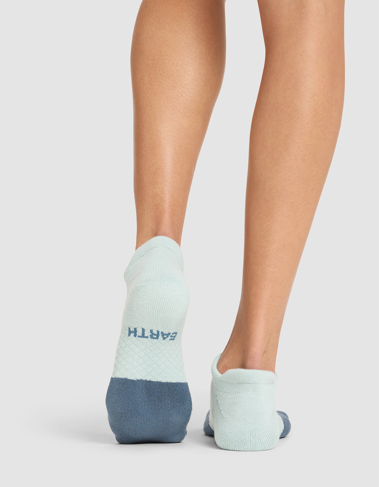 A person is shown from behind wearing Cozy Earth's Essential Ankle Socks in light and dark blue, with "EARTH" printed on the heel of the left sock, standing on a light-colored surface. 