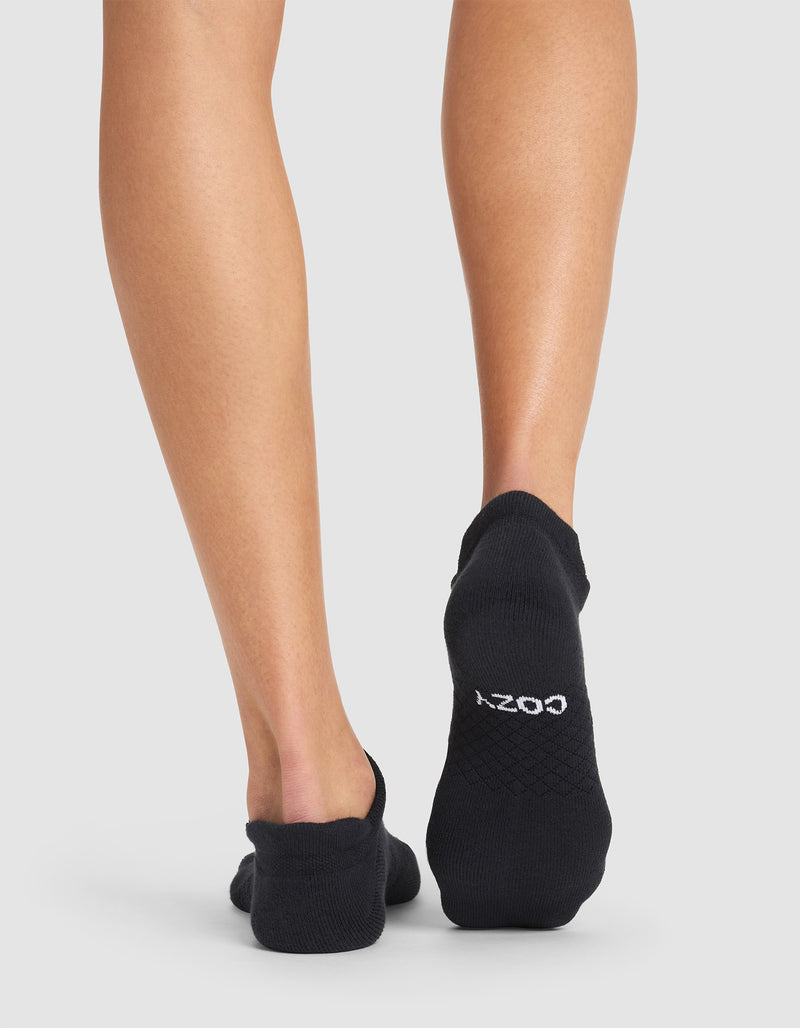 A person stands barefoot on a white background, wearing black Essential Ankle Socks by Cozy Earth. The back of their legs and feet are visible, with "COZY" printed in white on the sole of one sock.