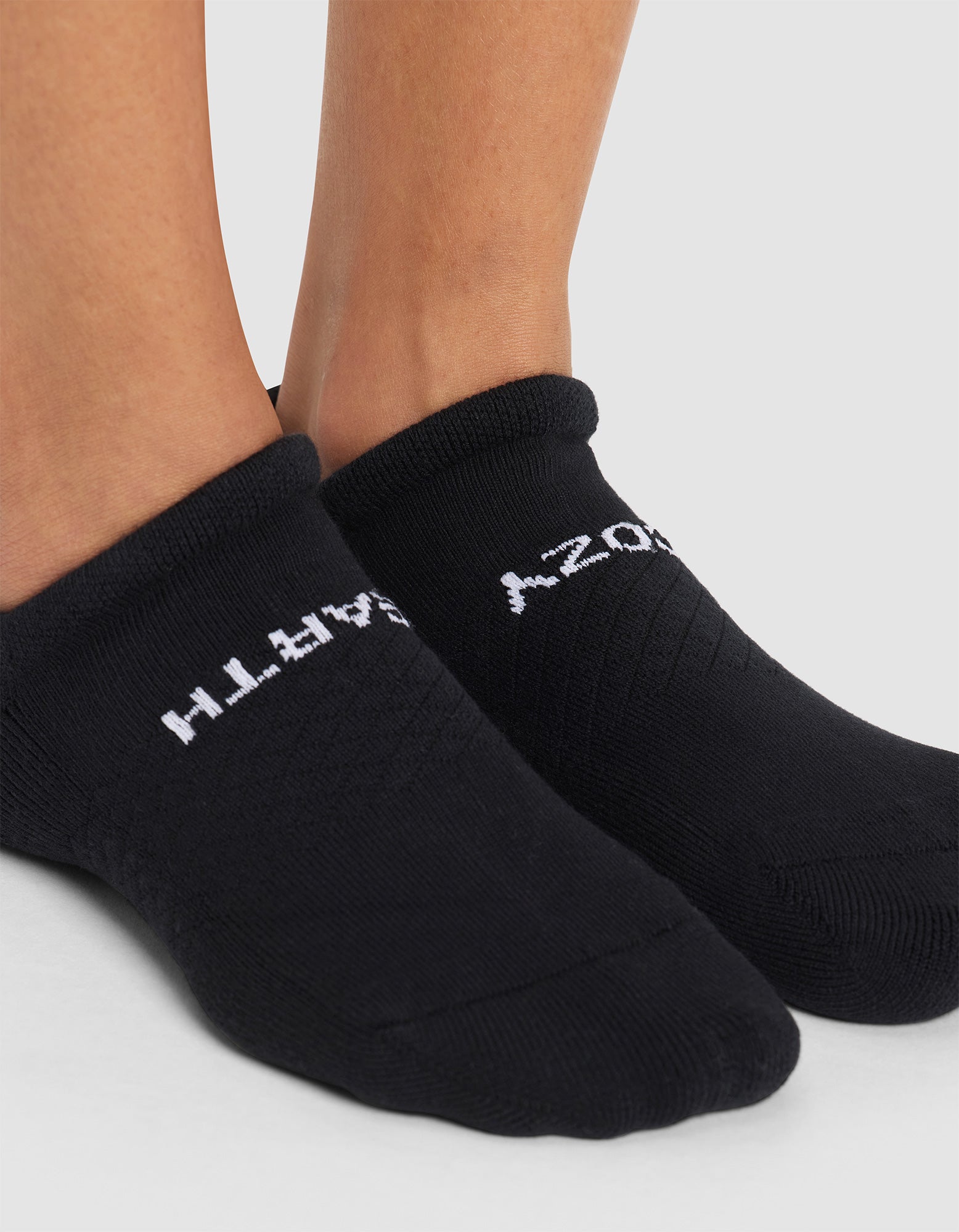 A close-up of two feet wearing Cozy Earth Essential Ankle Socks, black with white text on top, standing on a light gray surface. The sock text is partially visible. 