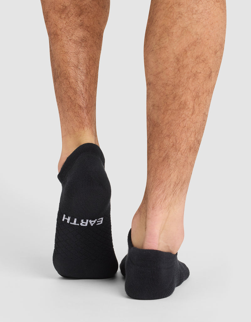 A close-up shows the back of a person's lower legs wearing Cozy Earth Essential Ankle Socks in black; "EARTH" is written in white on the left heel. The background is plain and light-colored.
