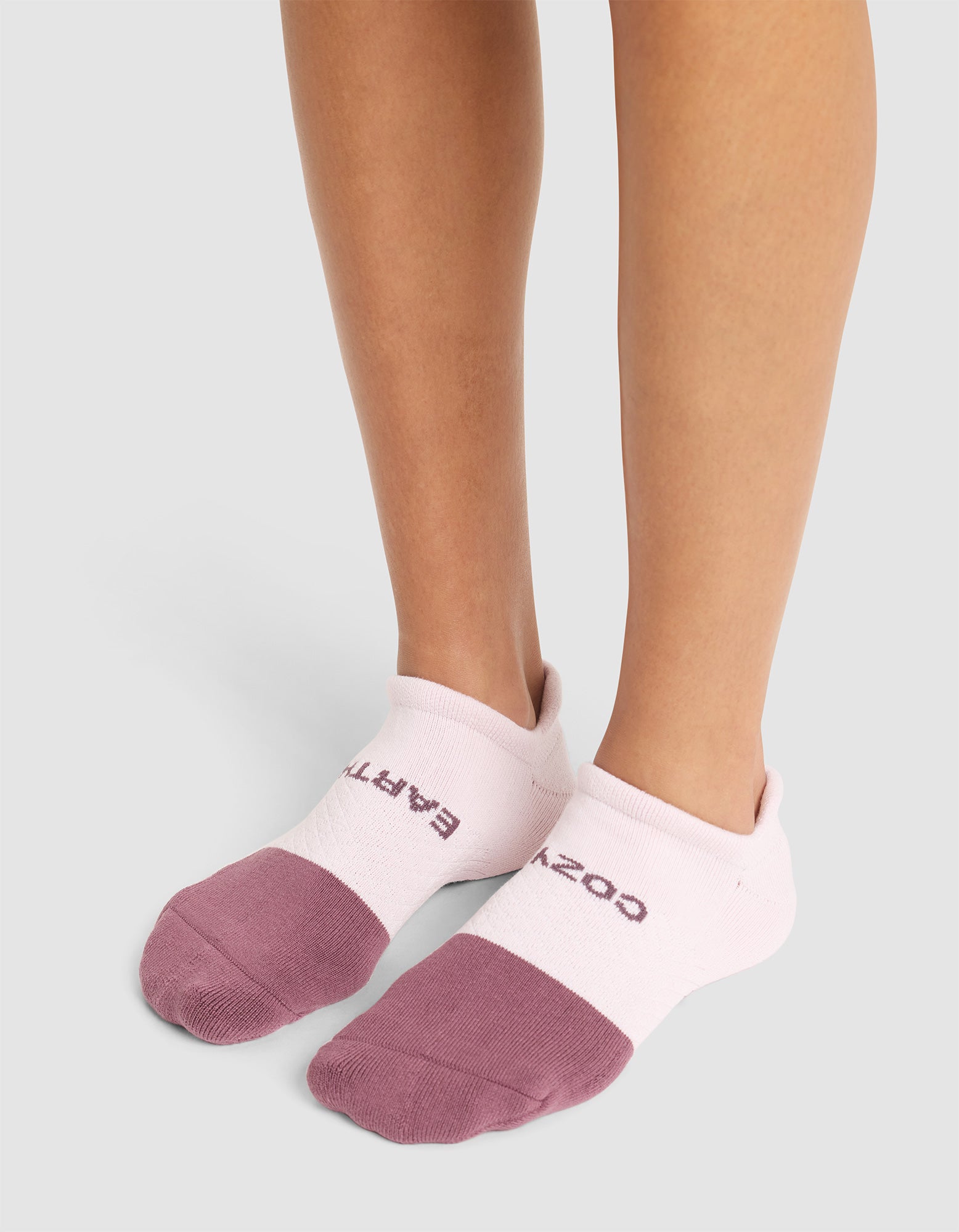 A person wearing Cozy Earth Essential Ankle Socks—white uppers with mauve toes and partial text near the cuffs—stands on a light gray surface. Only their lower legs and feet are visible. 