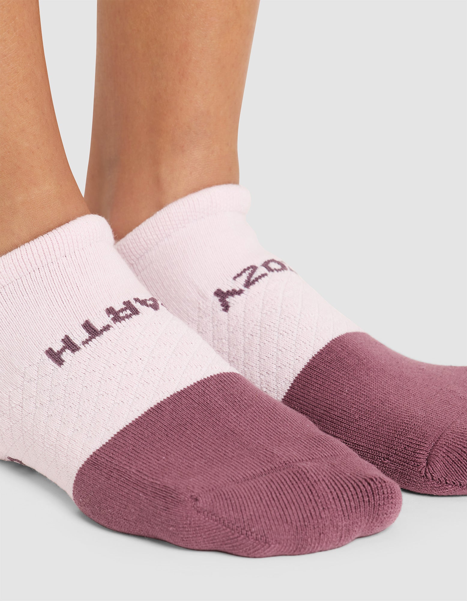 Close-up of two feet wearing Cozy Earth Essential Ankle Socks in pink and mauve, featuring a textured pattern with partially visible text across the top, set against a plain light gray background. 