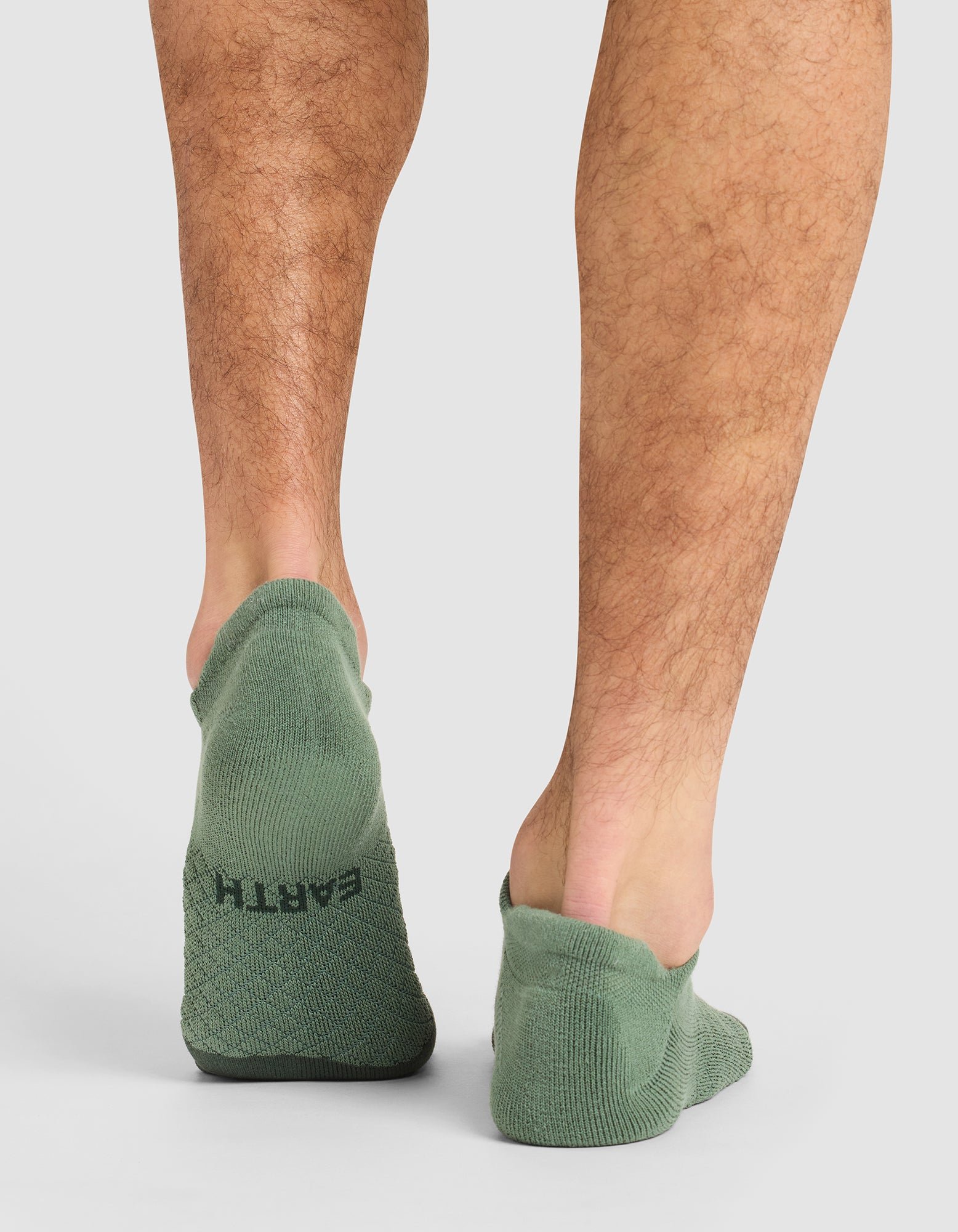 A person stands on a white surface, shown from the calves down, wearing green Cozy Earth Essential Ankle Socks. "EARTH" is printed in black on the heel of the left sock. 