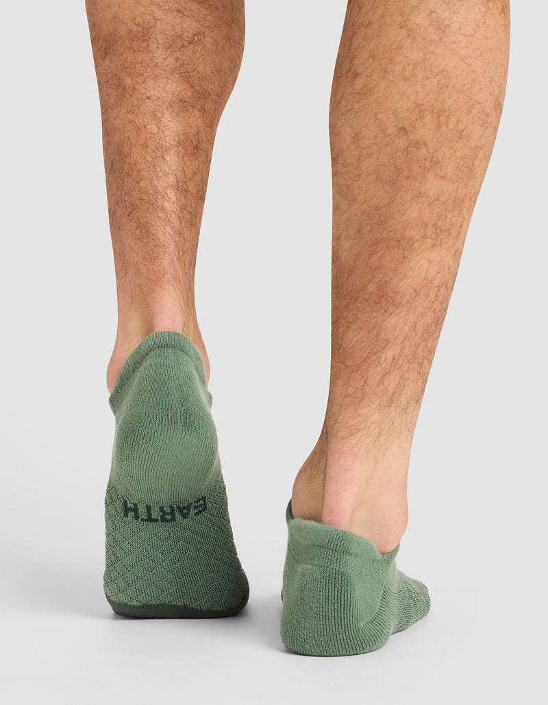 A person stands on a white surface, shown from the calves down, wearing green Cozy Earth Essential Ankle Socks. "EARTH" is printed in black on the heel of the left sock.
