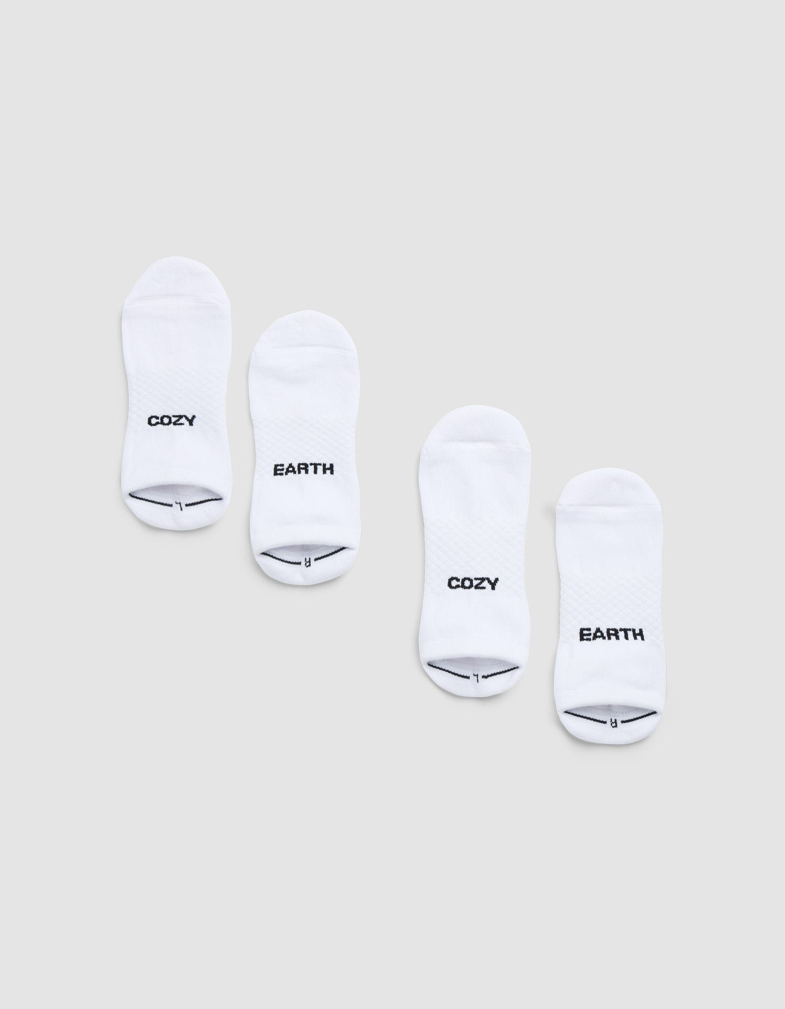 Two pairs of Cozy Earth Essential Ankle Socks are displayed side by side on a light gray background, each featuring black text near the toes—one in each pair reads "COZY" and the other reads "EARTH. |Color:White