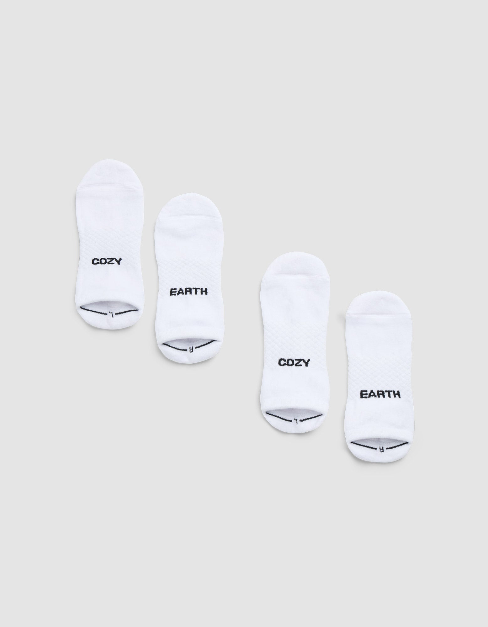 Two pairs of Cozy Earth Essential Ankle Socks are displayed side by side on a light gray background, each featuring black text near the toes—one in each pair reads "COZY" and the other reads "EARTH. |Color:White