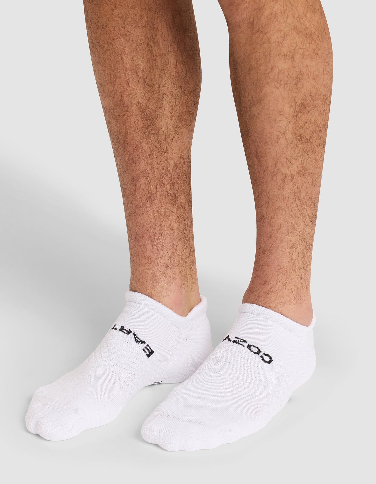 A close-up of a person's lower legs wearing Cozy Earth Essential Ankle Socks in white with black text, standing on a neutral gray background. 