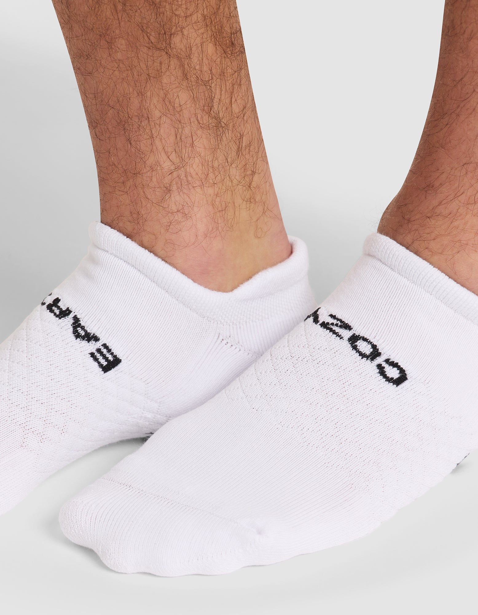 Close-up of a person's lower legs and feet wearing Cozy Earth's Essential Ankle Sock in white, featuring black text "EAR" on one sock and "OZ" on the other. The background is a plain light gray. 