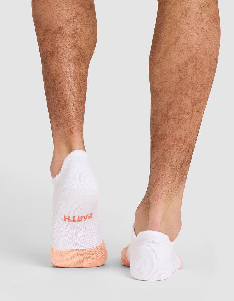 Someone stands on a light gray surface wearing Cozy Earth Essential Ankle Socks—white with light orange accents. "EARTH" appears in orange lettering on the back of the left sock. Only their lower legs and feet are shown.