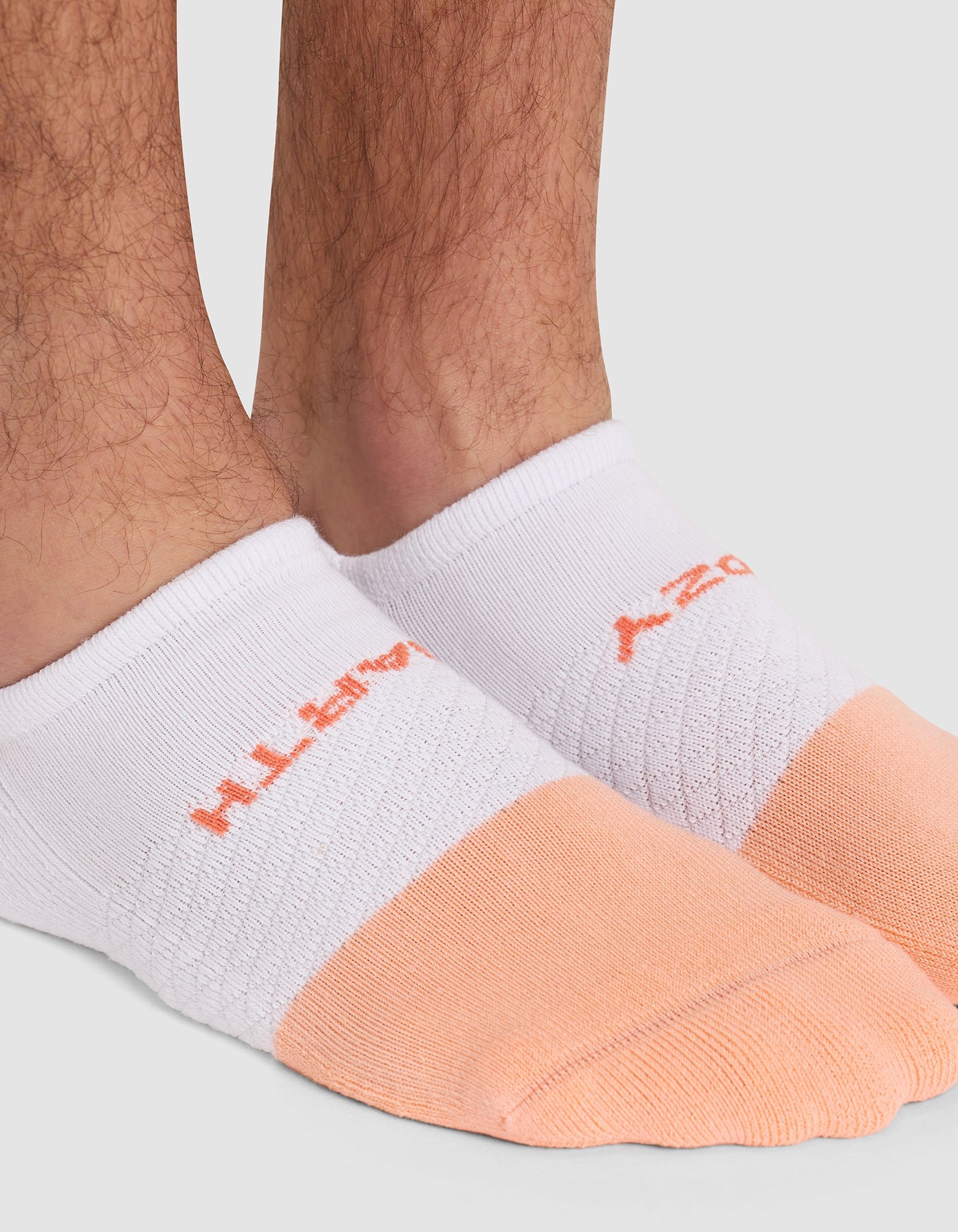 Close-up of a person's lower legs and feet wearing Cozy Earth Essential Ankle Socks in white and light orange, featuring orange text on top. Background is plain light gray. 