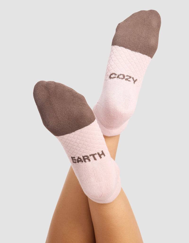 A woman wears Cozy Earth's Women's Essential Ankle Socks in pink with brown toes and heels. "EARTH" is on the left sock, "COZY" on the right. The background is plain light gray.