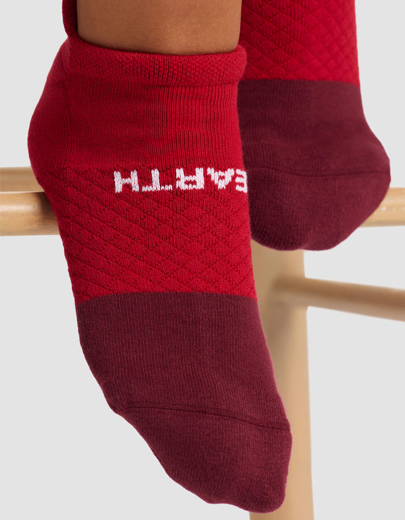 Close-up of feet in Cozy Earth's Women's Essential Ankle Sock—red with darker red toes and heels. "EARTH" in white appears upside down on the sole. The feet rest on a wooden bar.