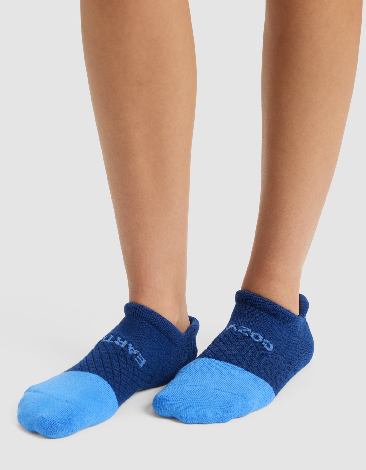 A person stands on a white surface wearing Cozy Earth Essential Ankle Socks in blue with darker blue cuffs and accents; "EARTH" appears on the left sock and "GOOD" on the right. 