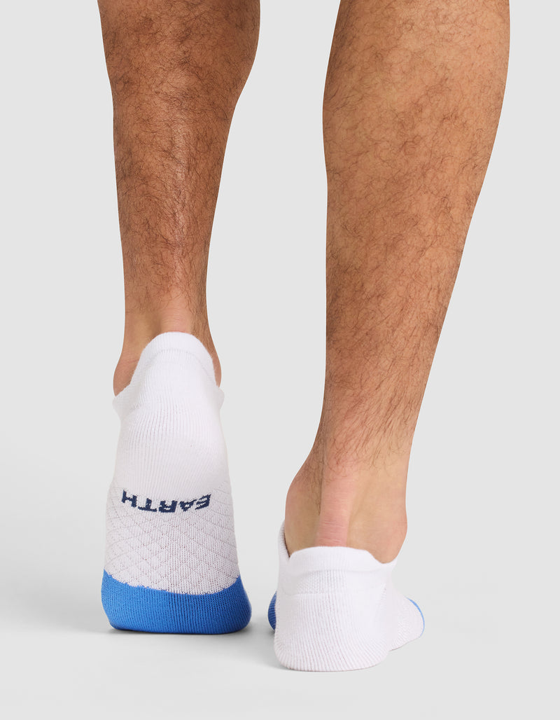 A person stands on a light surface, seen from behind, wearing Cozy Earth Essential Ankle Socks in white with blue heels. Only their lower legs and feet are visible, and "EARTH" appears on the left sock.