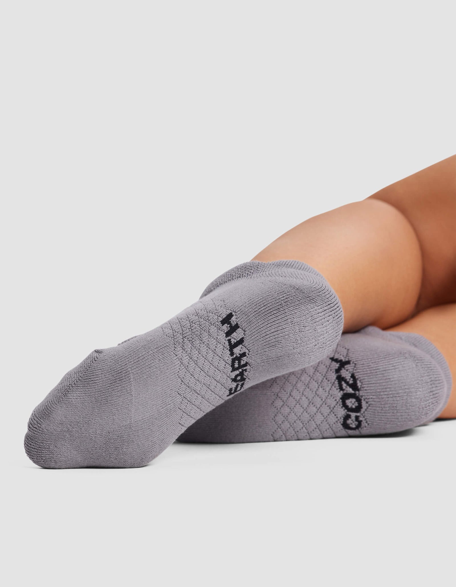 Close-up of a person lying down with feet facing the camera, wearing Cozy Earth Women's Essential Ankle Socks in light gray, featuring "COZY" on one and "CRAFTY" on the other in black letters.