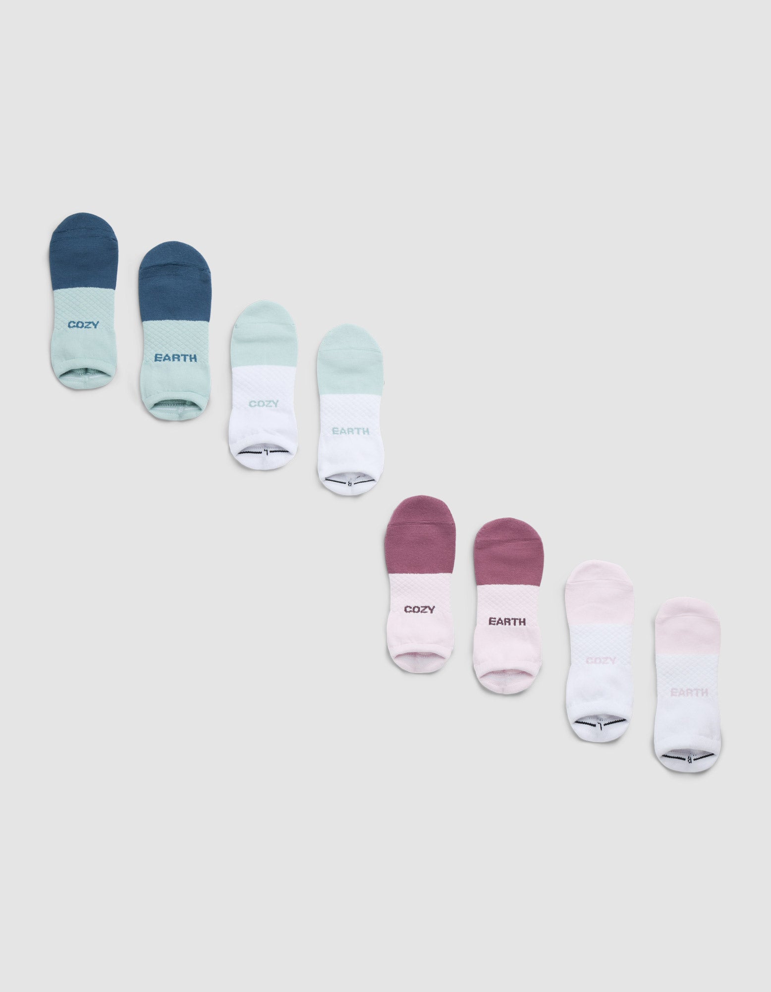 A Cozy Earth Essential Ankle Sock 4-Pack is shown in shades of blue, green, white, pink, and purple on a light gray background. Some socks have "COZY" or "EARTH" printed near the toes. 