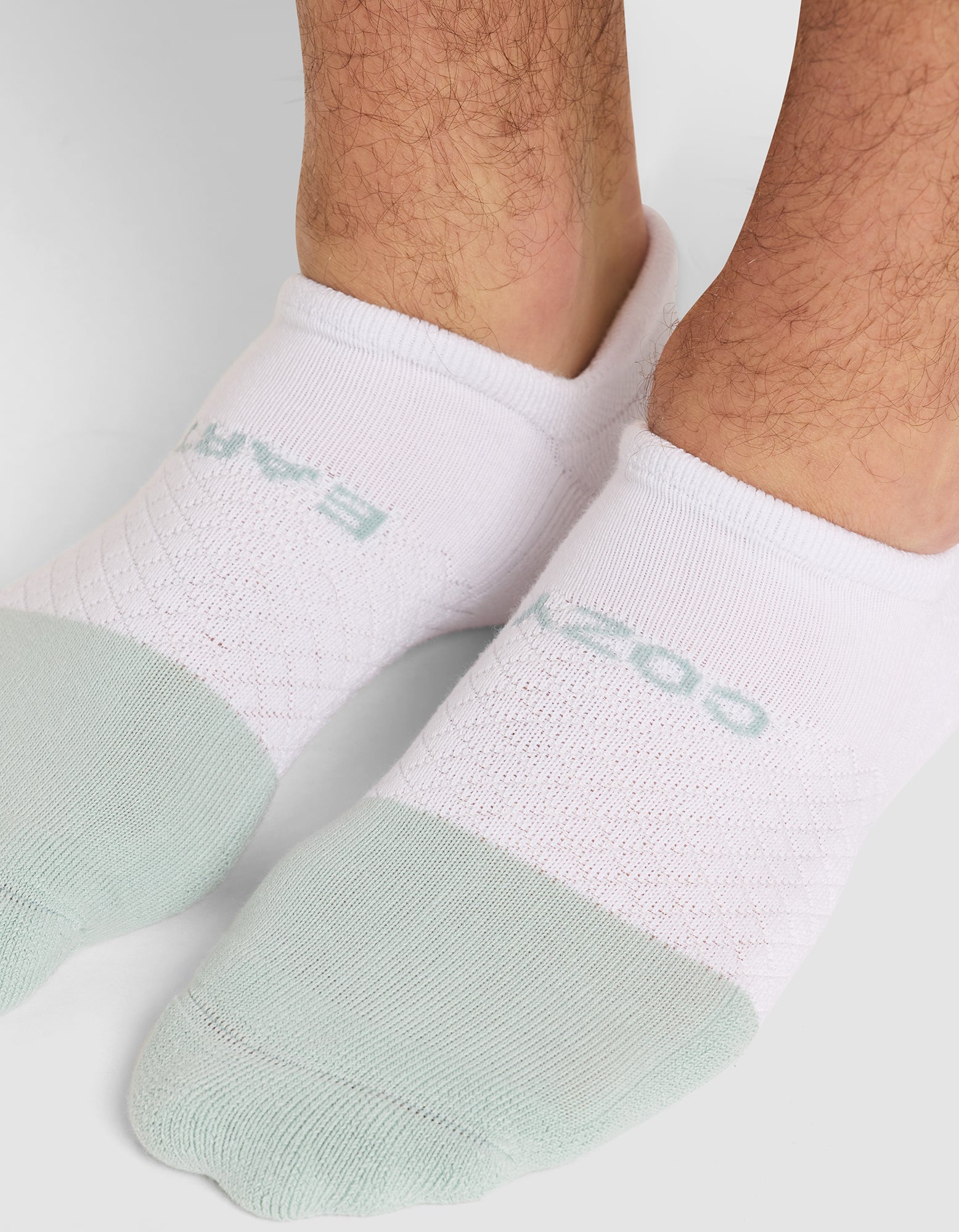 Close-up of a person's lower legs and feet wearing Cozy Earth Essential Ankle Socks with light green toes and light blue text on top, standing on a white surface. 