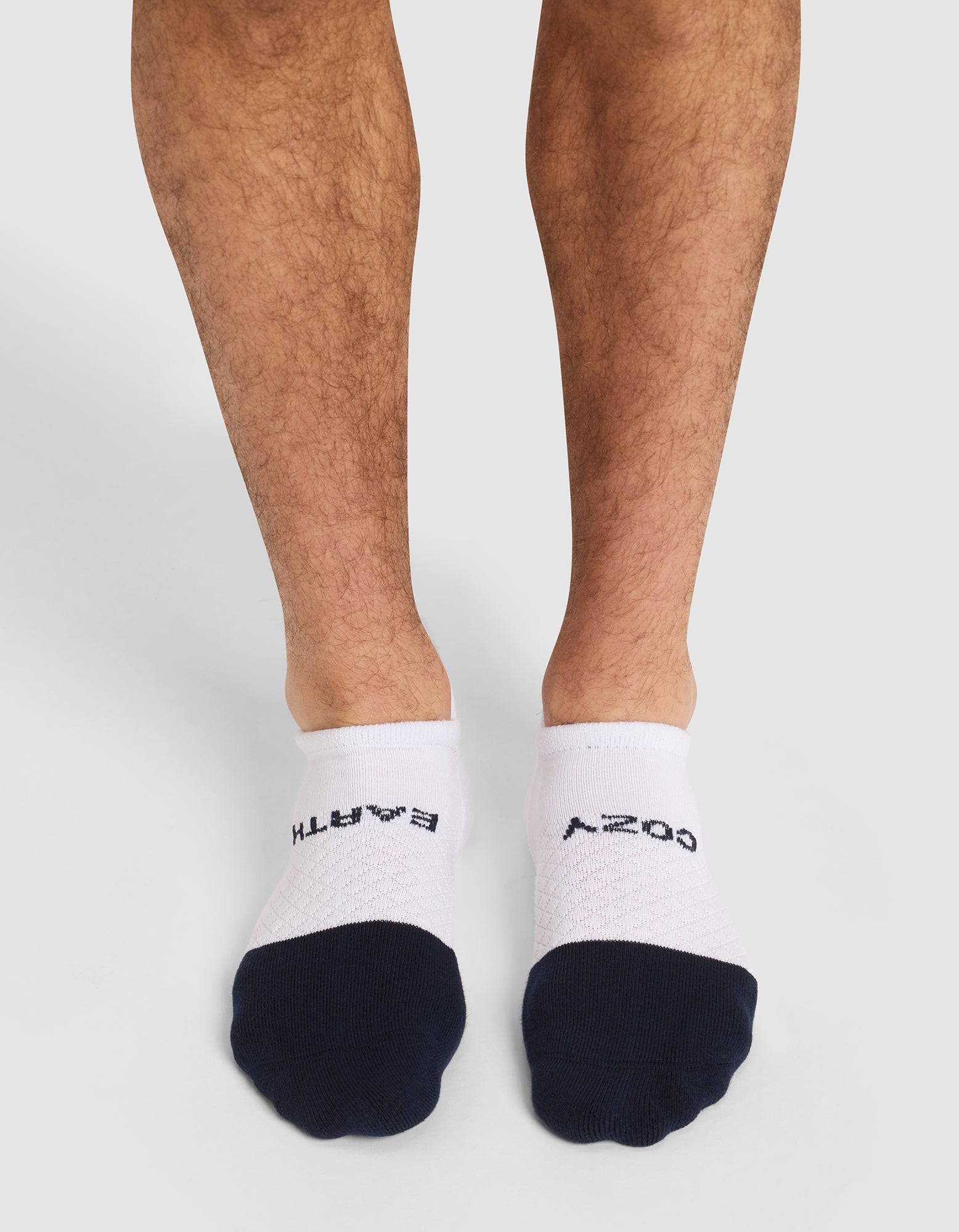 A person stands on a white background wearing Cozy Earth Essential Ankle Socks—white no-show socks with black toes; "BATH" is written on the left sock and "COZY" on the right in black letters. 