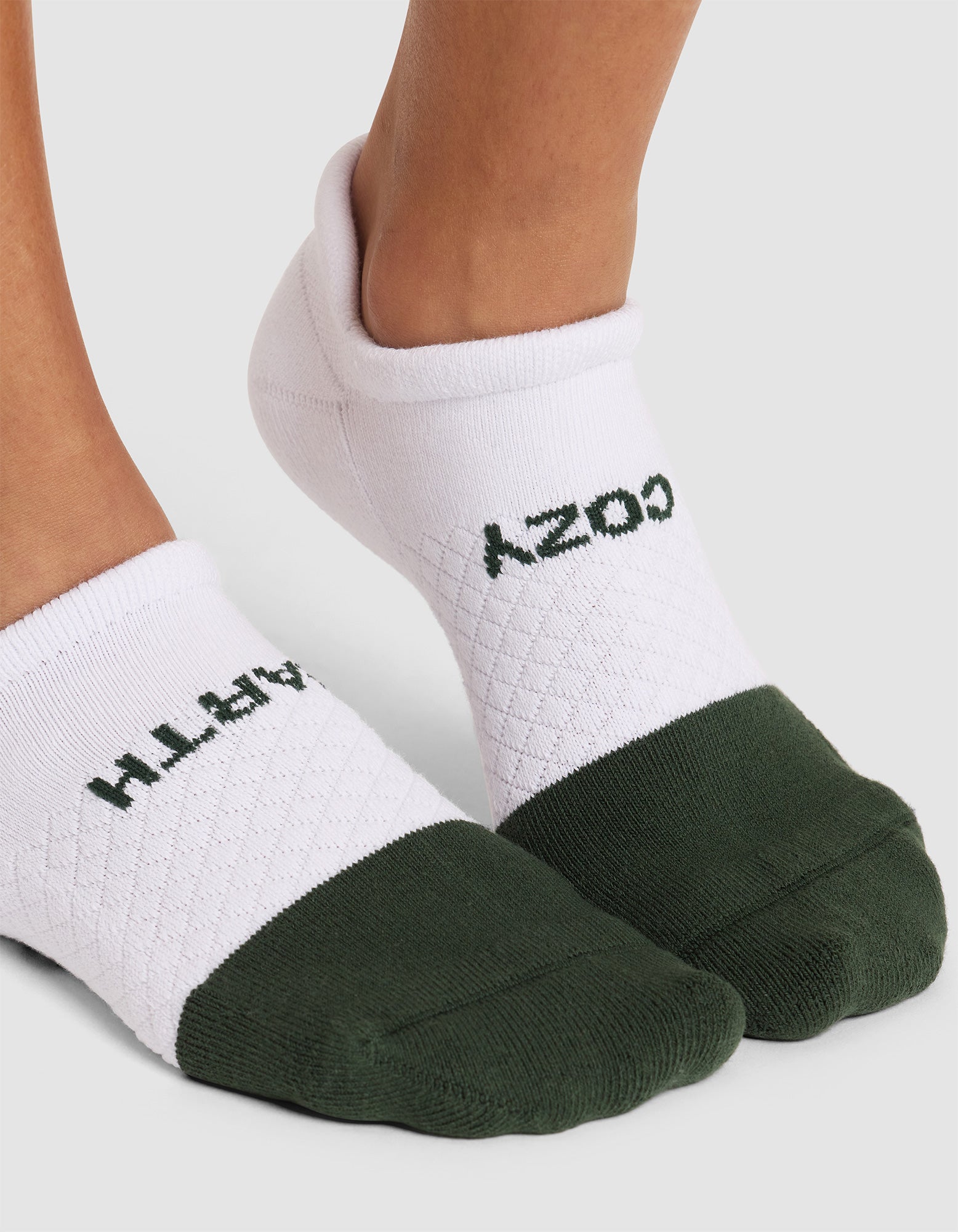 A close-up of feet in Cozy Earth Essential Ankle Socks, featuring a white and dark green design, quilted texture on top, and the word “COZY” printed in black, shown upside down from this angle. 