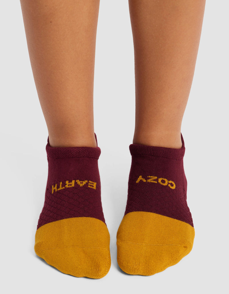 A person wears Cozy Earth’s Women's Essential Ankle Socks—maroon with yellow toes and upside-down "COZY" and "EARTH" text—standing on a light surface.