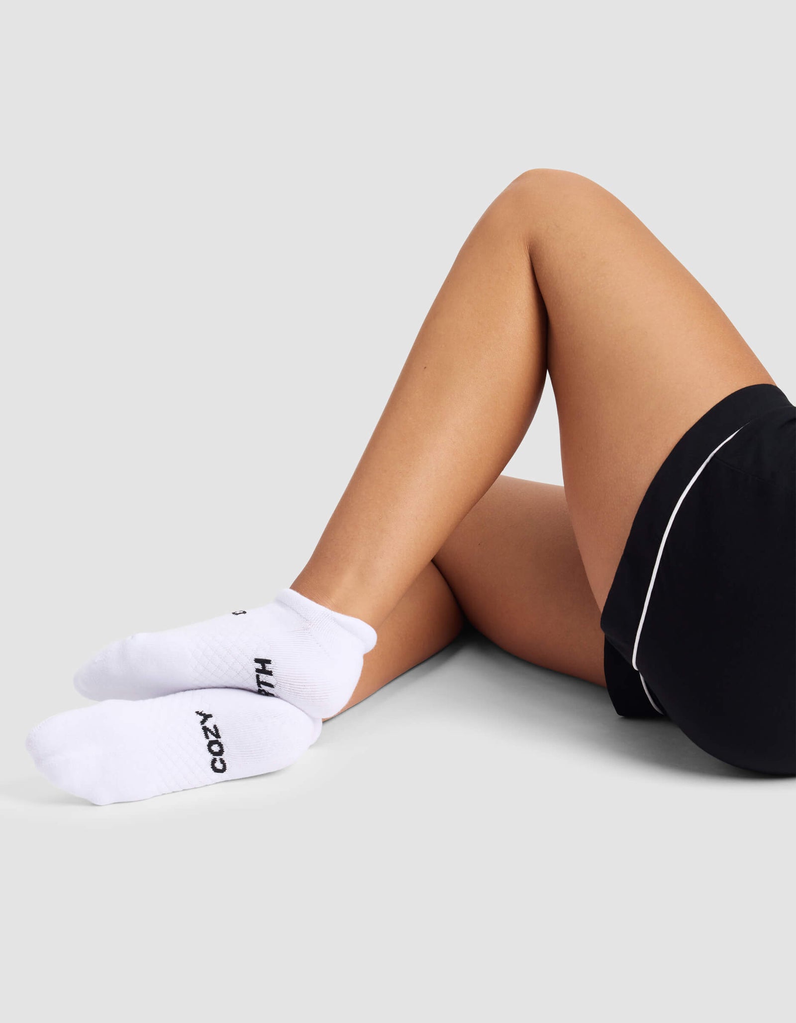 A woman wears Cozy Earth Women's Essential Ankle Socks with black text, paired with black shorts, sitting cross-legged on the floor against a light grey background.
