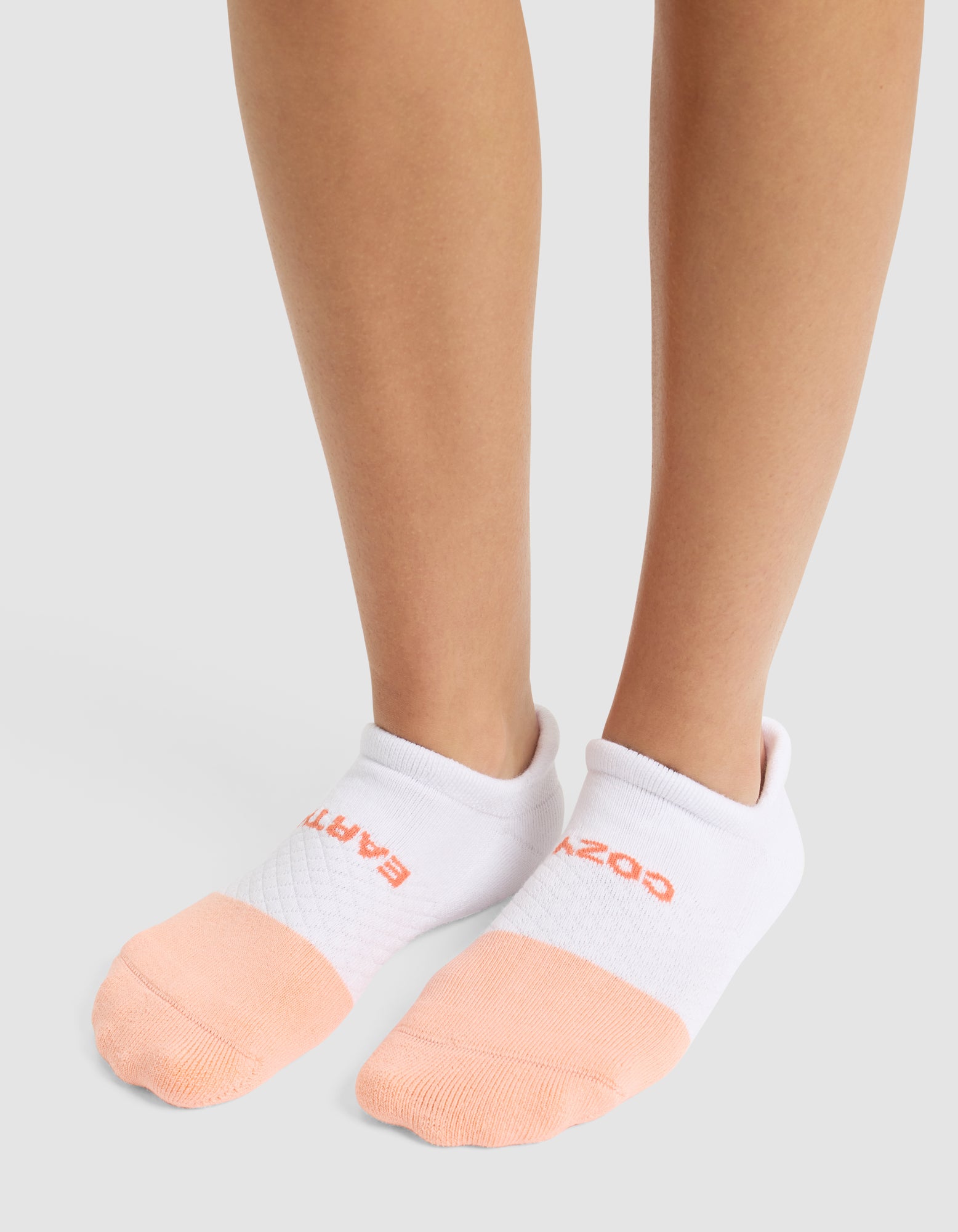 A person wearing Cozy Earth's Essential Ankle Sock—featuring white tops, peach toes, and red text near the cuff—stands on a light gray surface. 