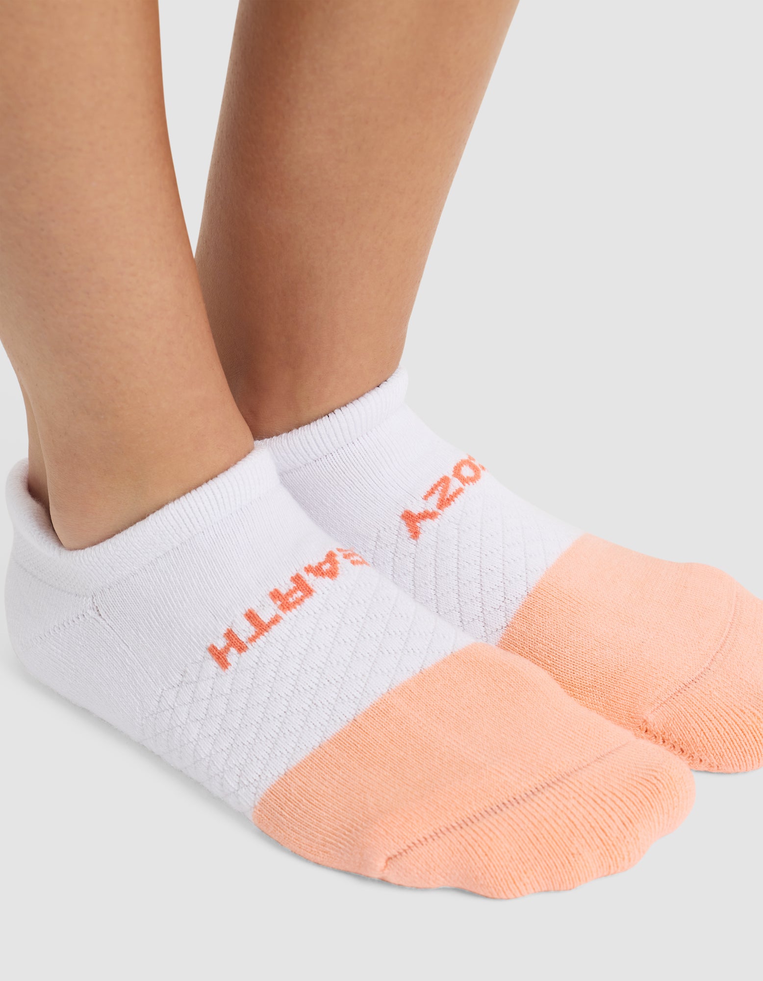 Close-up of a person's feet wearing Cozy Earth's Essential Ankle Sock in white, featuring peach-colored toes and heels. "EARTH" is stitched in orange on the side. 
