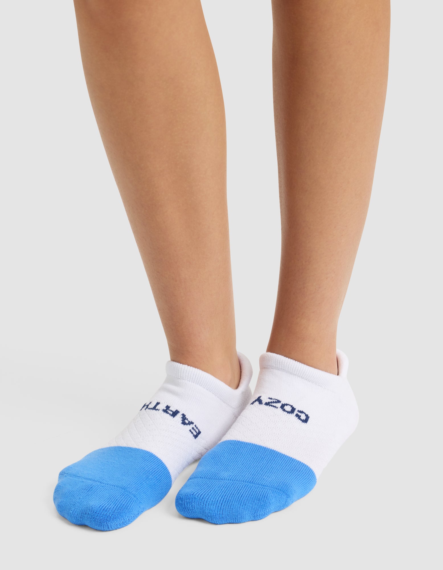 A person wears Cozy Earth Essential Ankle Socks—white with blue toes. "EARTH" is printed in blue on the left sock and "COZY" on the right. Both feet are standing on a light surface. 