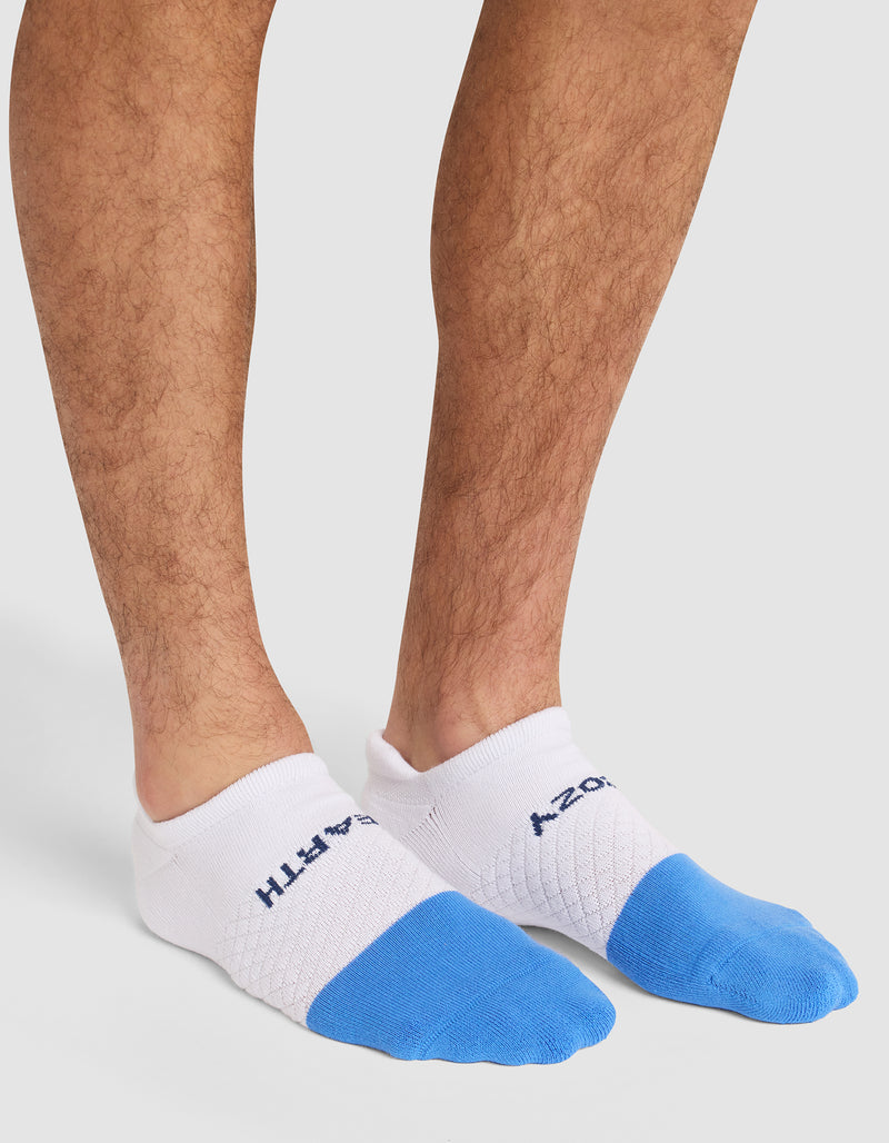 A person wears Cozy Earth Essential Ankle Socks featuring white heels, blue toes, text, and a mesh pattern on top, standing on a light surface.