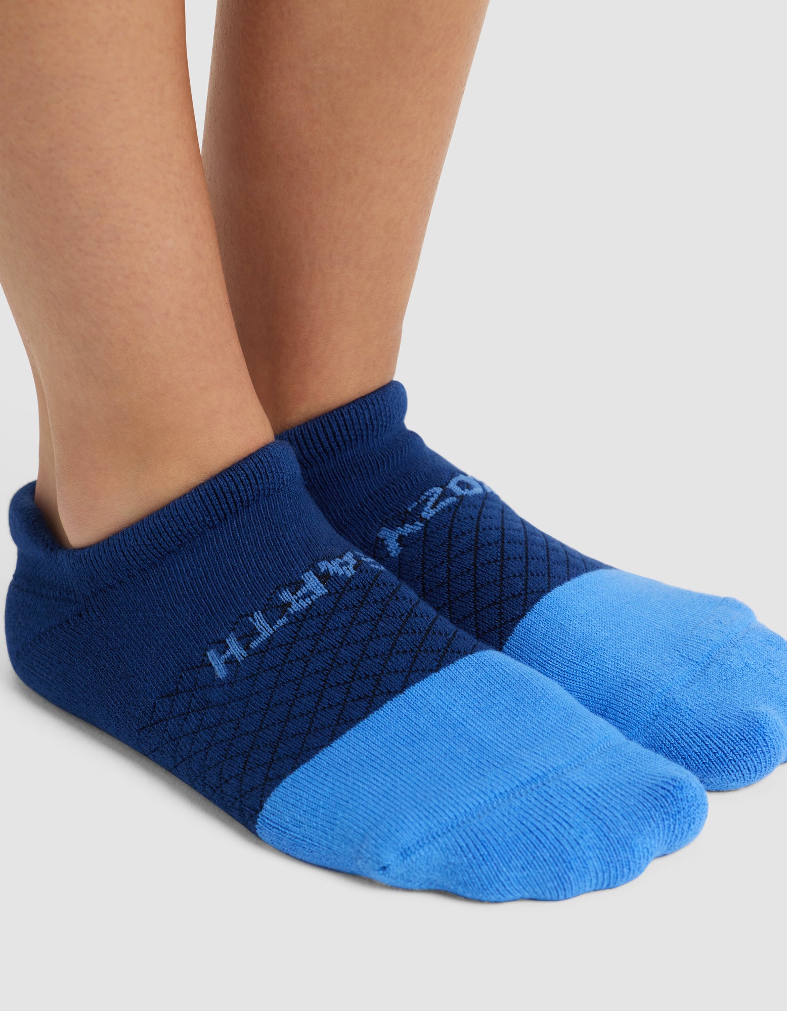 A pair of bare feet wears Cozy Earth's Essential Ankle Sock in blue, featuring a darker blue pattern, “WITH 202” text on top, and a textured design, all set against a plain background. 