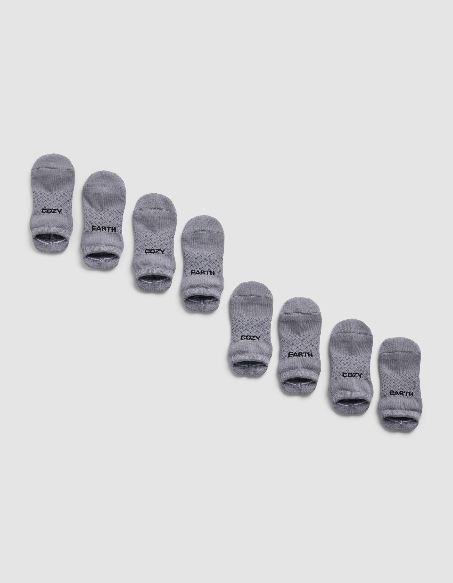 Four pairs of gray Essential Ankle Socks from Cozy Earth are arranged diagonally on a light gray background, each featuring "COZY EARTH" printed in black near the toes.|Color:Stone