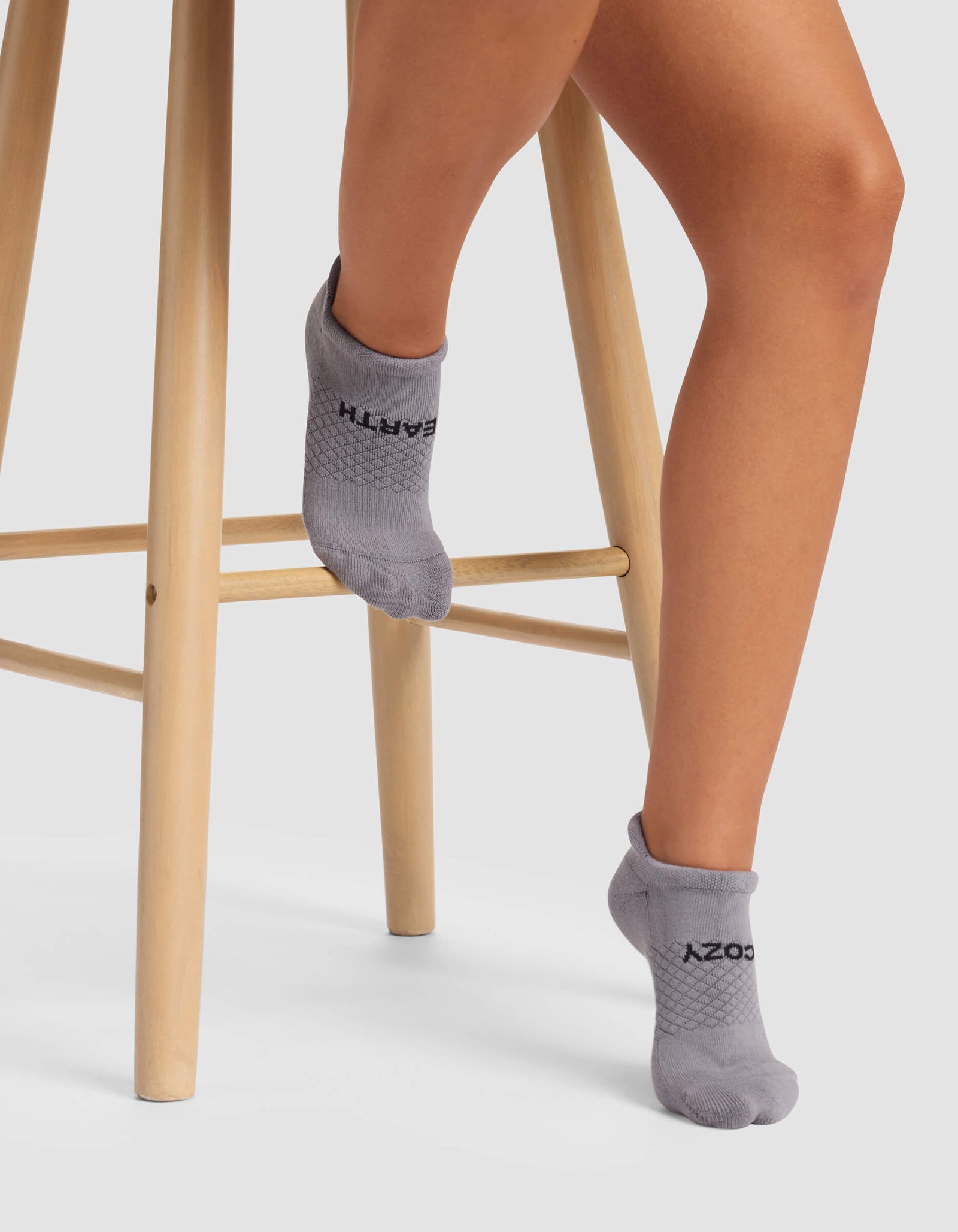 A person wearing Cozy Earth Women's Essential Ankle Socks in gray sits on a light wooden stool, with one foot on a lower rung and the other pointed downward.