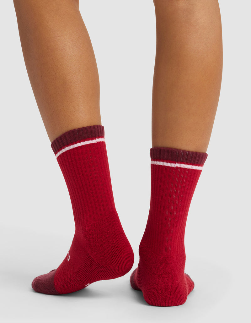 A person stands with their back to the camera, wearing Cozy Earth Essential Calf Socks—red with dark maroon cuffs and a white stripe—on a light background.