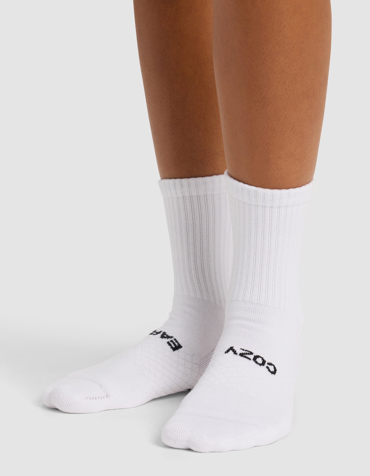 A person wearing Cozy Earth’s Essential Calf Sock—white ribbed crew socks with black text reading "COZY" on one and "BABY" on the other—stands on a light surface.