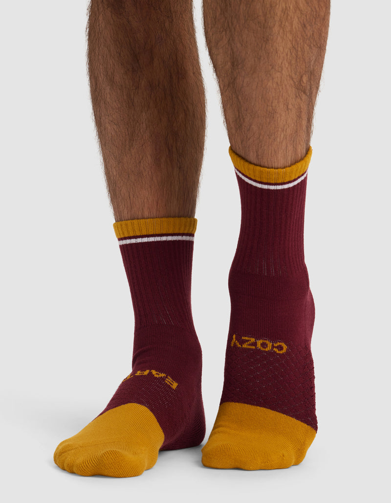 A person wears mismatched Cozy Earth Essential Calf Socks—both maroon with yellow toes and cuffs, but each features different yellow accents and text—shown standing on a white background.