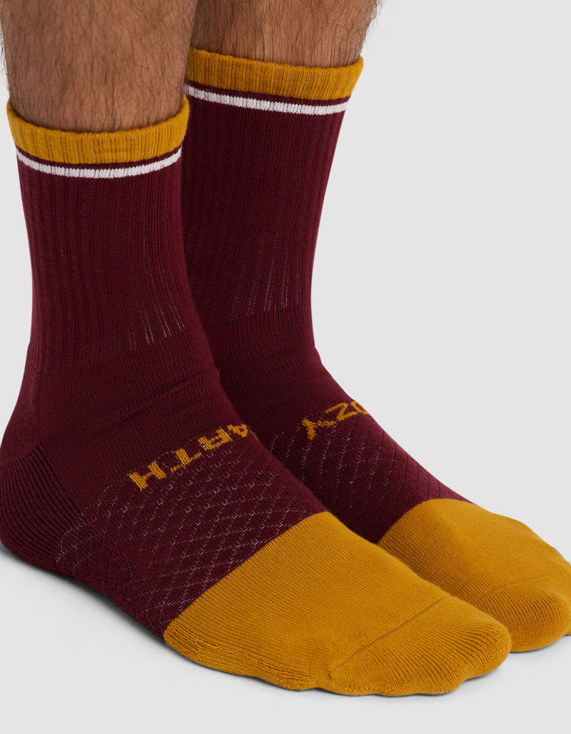 Two feet wear Cozy Earth's Essential Calf Socks in maroon with mustard yellow toes, heels, and cuffs, plus a white stripe near the top. “EARTH” is printed in yellow on one sock, “COZY” on the other.