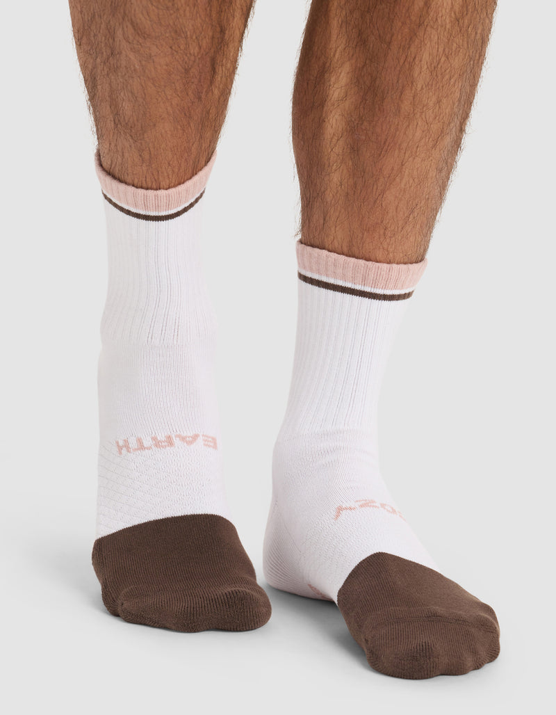 A person stands on a light gray surface wearing Cozy Earth Essential Calf Socks in white, featuring brown toes and a brown stripe, with "EARTH" faintly visible in pink on the left sock.