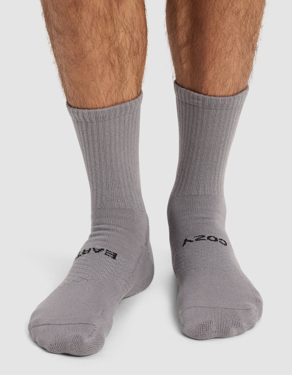 A pair of feet wearing Cozy Earth’s Men’s Essential Calf Sock in gray with black text on the toes stands on a white surface; the mid-calf socks are modeled by someone with light brown leg hair.