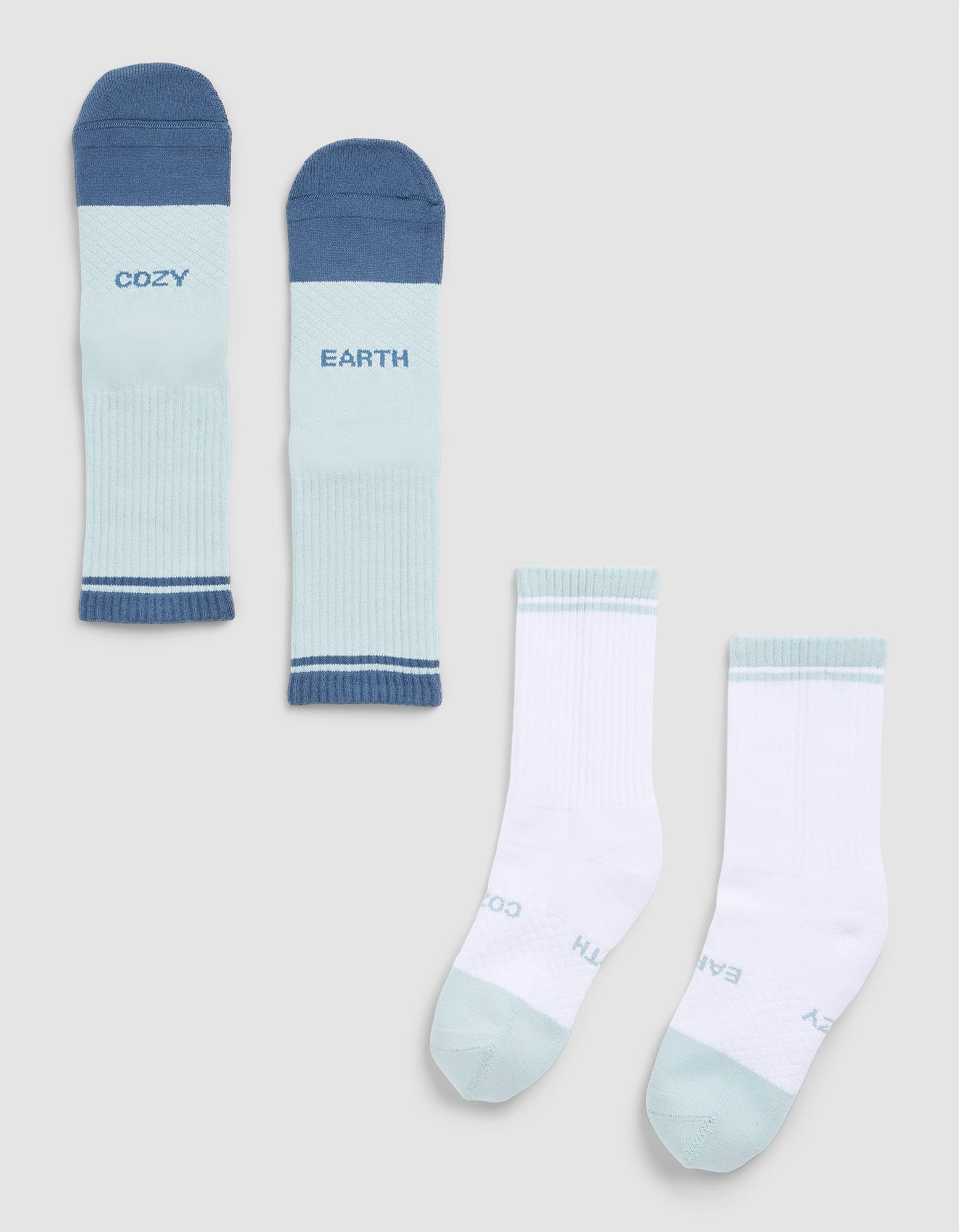 Two pairs of Cozy Earth Essential Calf Socks are displayed: one in blue tones with "COZY" and "EARTH" text, and another in white with light blue accents and similar branding, both arranged neatly on a light gray background. 