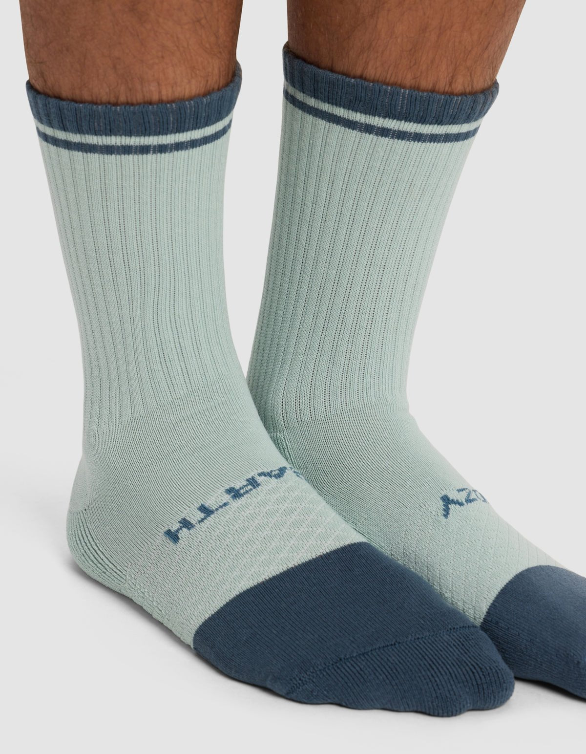 A close-up of lower legs wearing Cozy Earth's Essential Calf Sock - Last Chance: light blue crew socks with dark blue toes, heels, and cuffs, thin dark blue stripes near the top, and text printed on the tops of the feet.