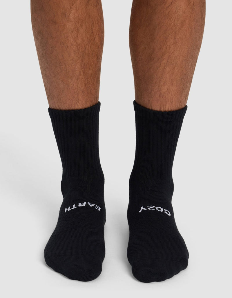 A person wears Cozy Earth Essential Calf Socks in black, featuring "EARTH" on the left and "COZY" on the right sock. Only their lower legs and feet appear against a plain background.