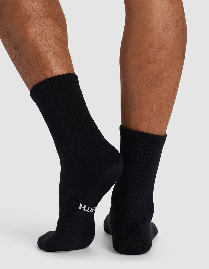 A close-up shows a person wearing Cozy Earth's Essential Calf Sock in black while standing on a light gray surface. "EARTH" is visible near the toes on one sock.