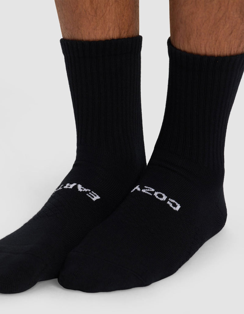 Close-up of lower legs and feet wearing Cozy Earth Essential Calf Socks in black. "EARTH" is printed on the left sock, "COSY" on the right. The plain, light background highlights the ribbed design.