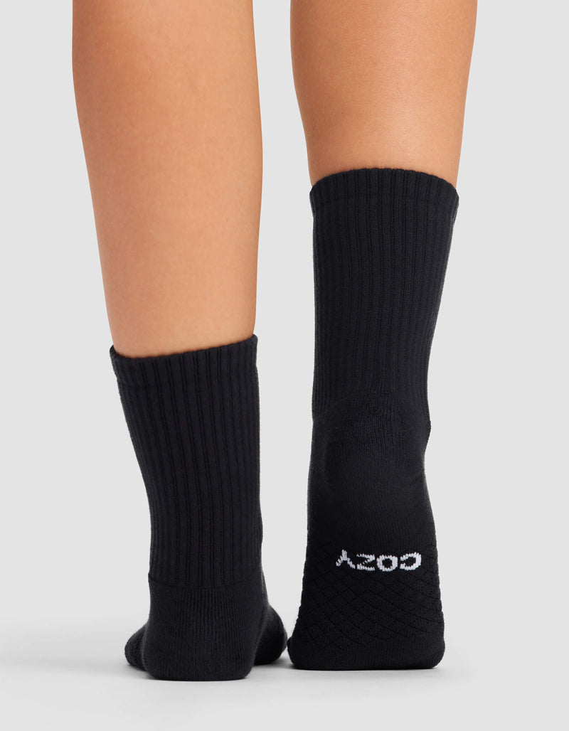 A person stands on a light surface wearing Cozy Earth's Women's Essential Calf Socks. The left sock is plain, while the right sole displays "COZY" in text.