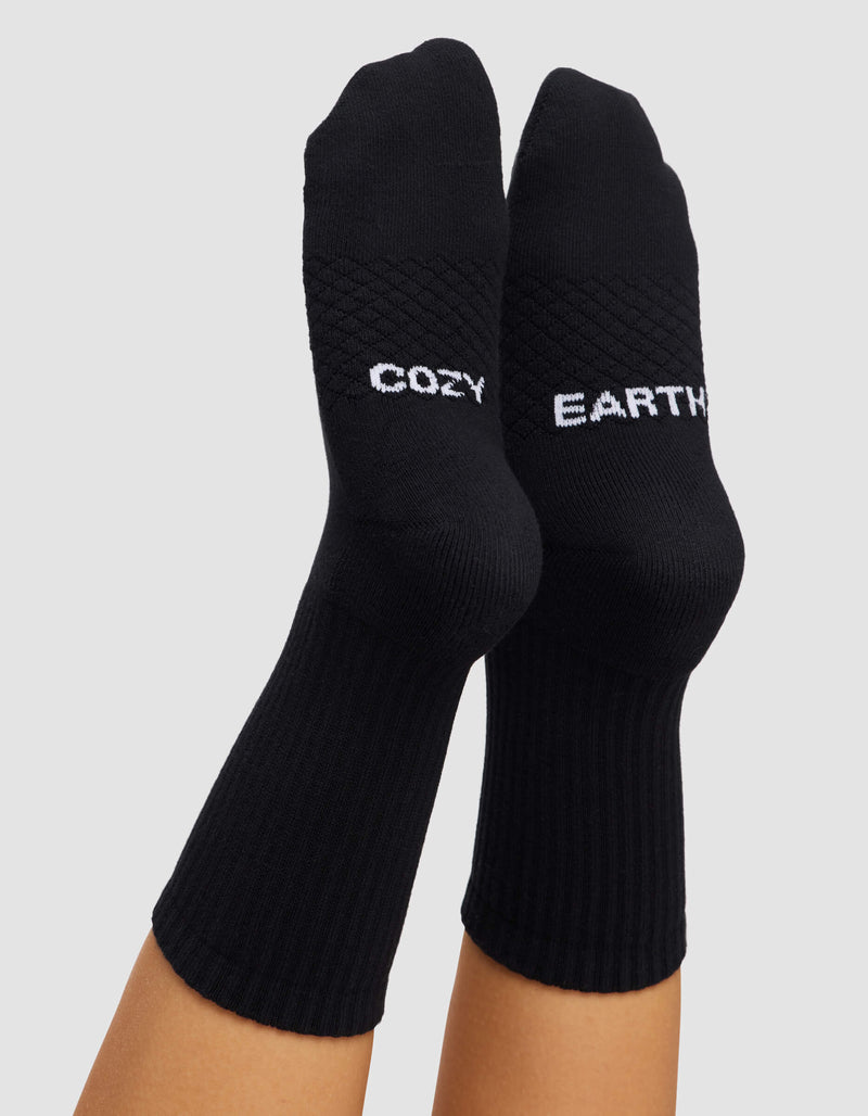 A pair of feet wearing Women's Essential Calf Sock by Cozy Earth, featuring “COZY” on one sock and “EARTH” on the other in light lettering, set against a plain light gray background.