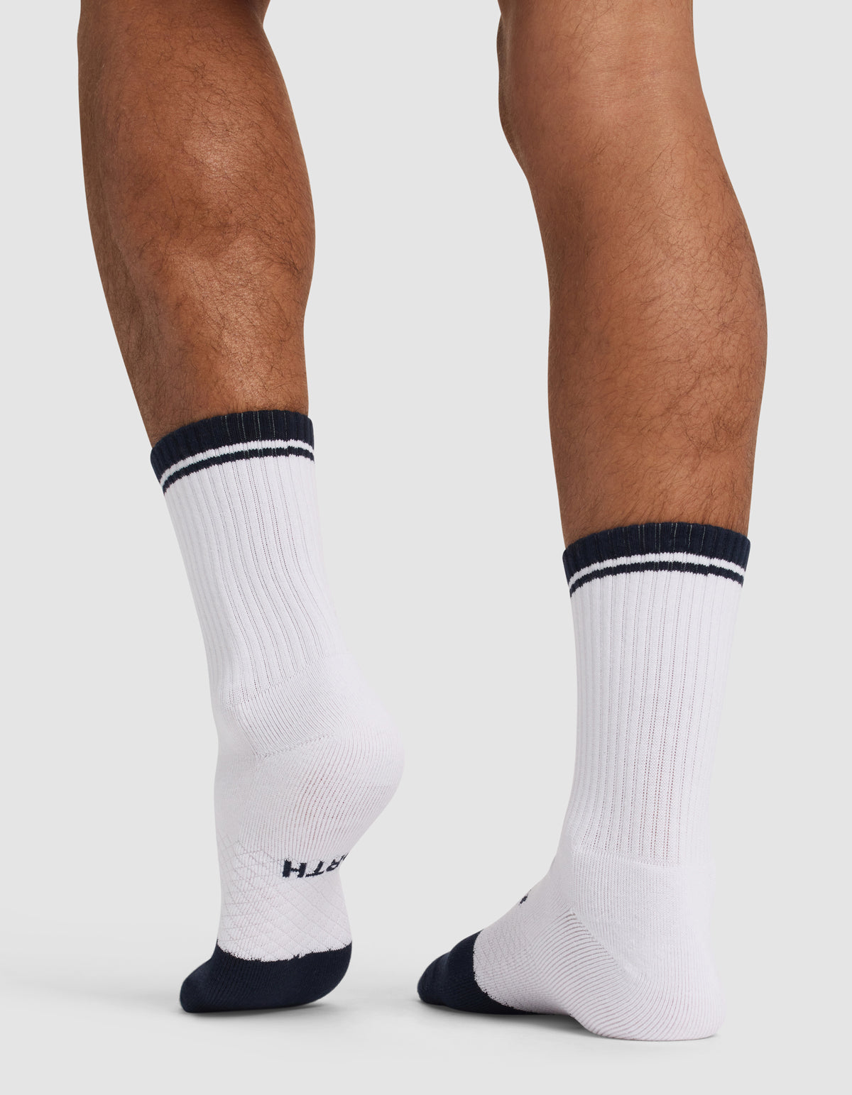 Someone stands on a light surface, viewed from behind with one heel slightly raised, wearing Cozy Earth's Essential Calf Sock - Last Chance in white with dark blue accents and stripes.
