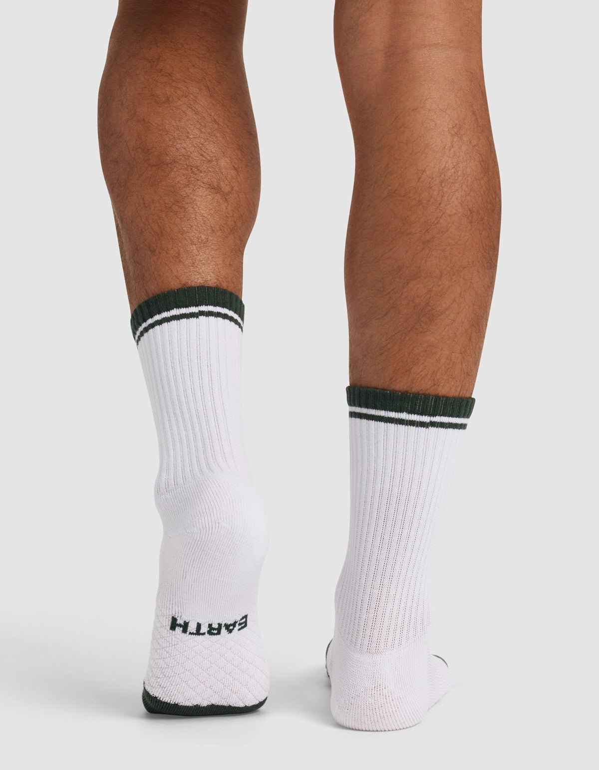 A person stands on a white surface wearing Cozy Earth's Essential Calf Sock – Last Chance, white with dark green stripes and "EARTH" visible on the sole. The photo is taken from behind.